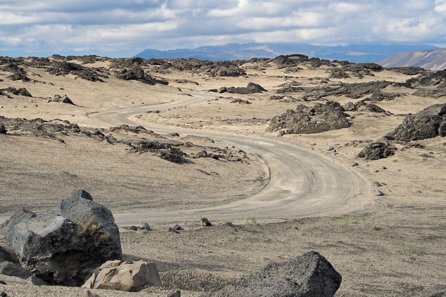 Eine sandige Straße führt durch eine karge Landschaft durchbrochen von schwarzen Felsen.