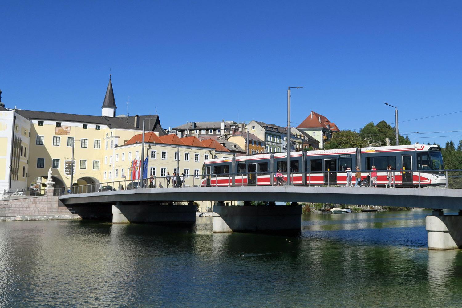 Eine Brücke überspannt einen Fluss, auf der Menschen zu Fuß laufen und eine S-Bahn fährt. Im Hintergrund sind Häuser und blauer Himmel.