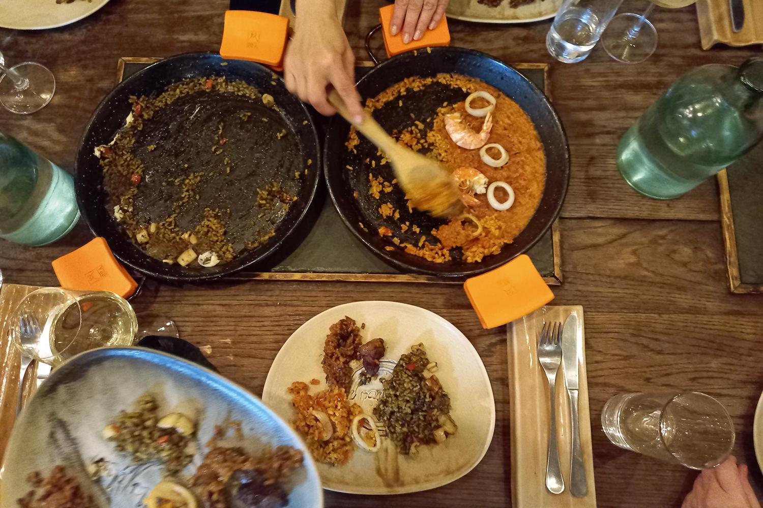 Ein gedeckter Tisch aus der Vogelperspektive, mittig sind zwei Pfannen platziert, eine ausgekratzte und eine noch halb gefüllt mit Paella