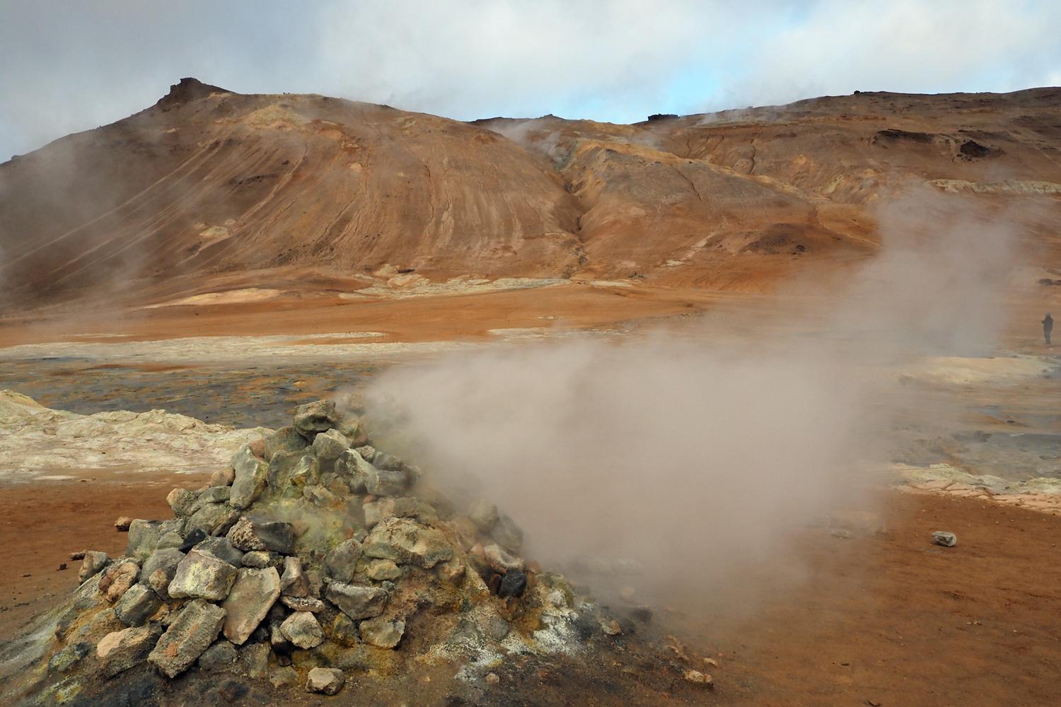 Das Bild zeigt einen dampfenden, schwefeligen Steinhaufen im Vordergrund mit rostfarbenen Hügeln im Hintergrund. 