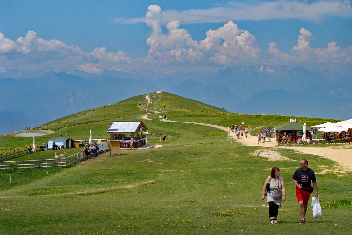 Grüne Bergwiese mit zwei Hütten und vereinzelt Menschen auf dem Weg