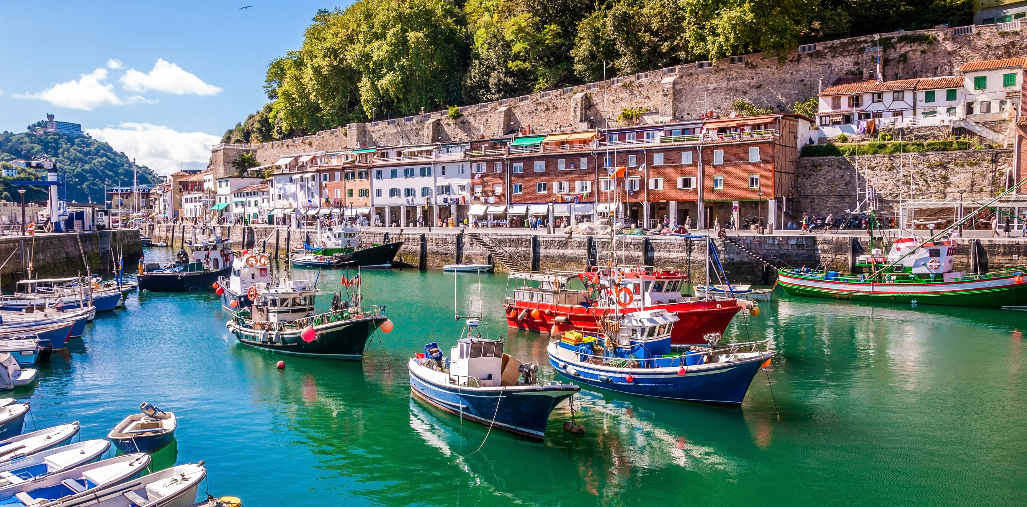 Hafen von Donostia / San Sebastián: bunte Fischerboote, alte Häuserzeile, blauer Himmel, im Hintergrund  ein Hügel mit Burg