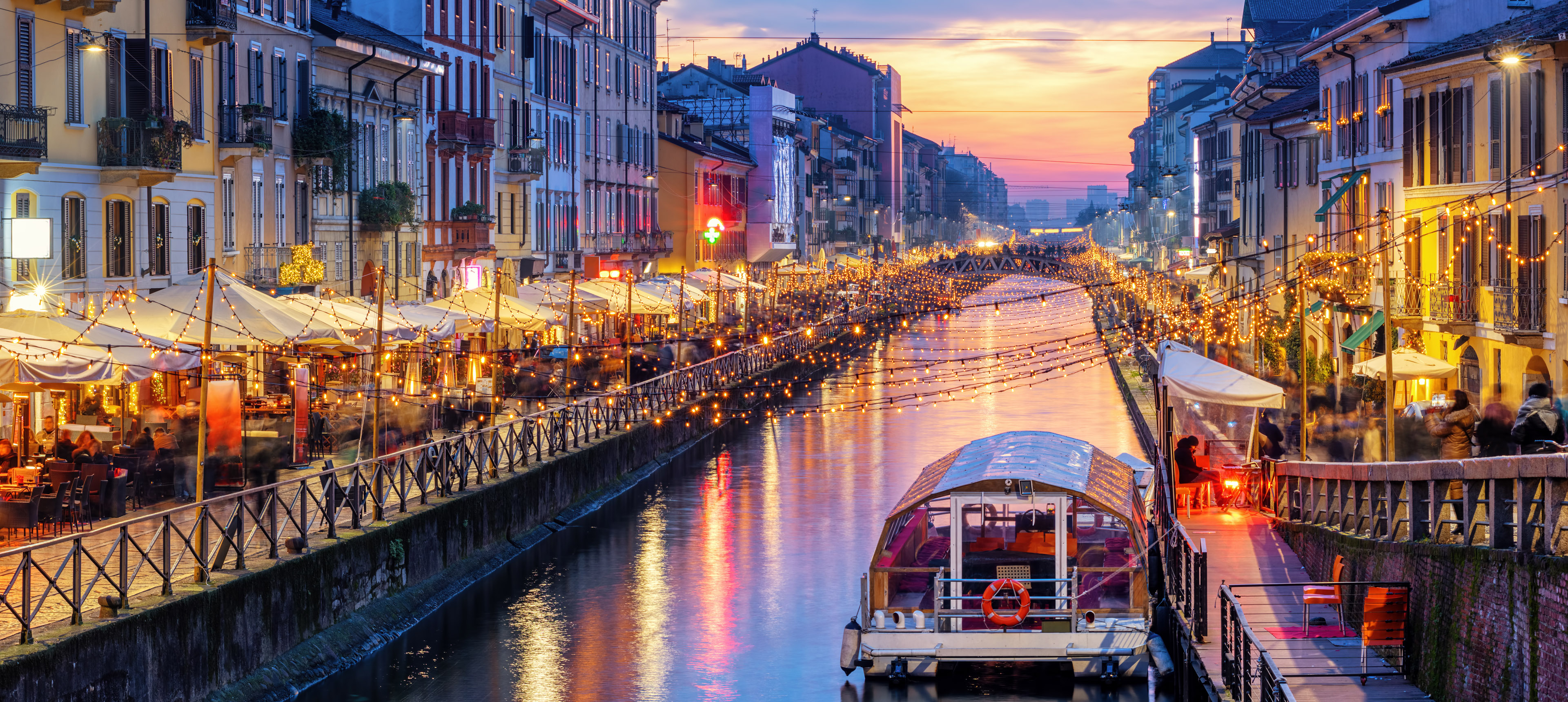 Der mit vielen kleinen Lichtern beleuchtete Kanal Naviglio Grande in Mailand bei Sonnenuntergang, im Vordergrund ein Passagierboot, links und rechts alte Häuser mit Terrassencafés