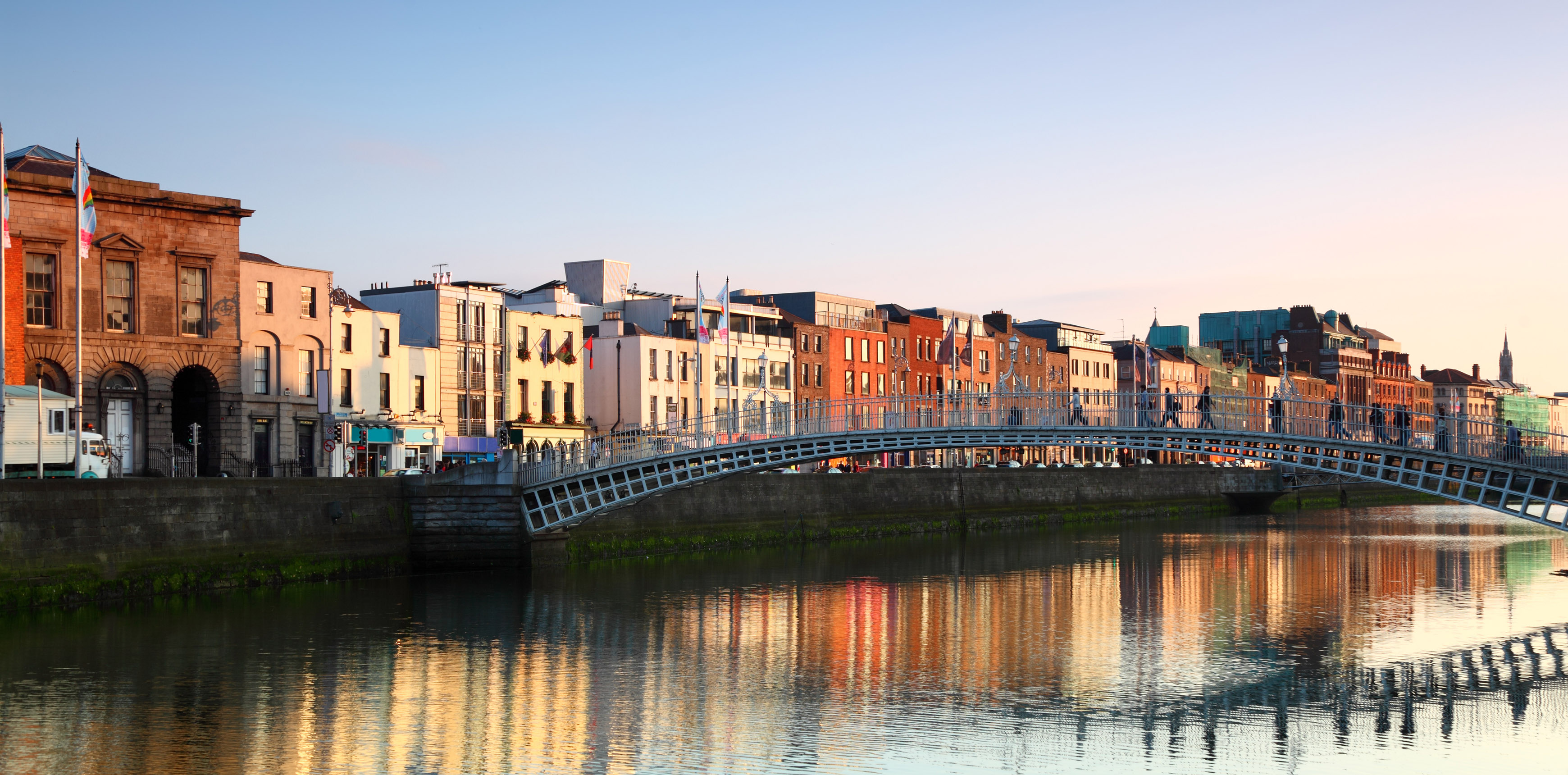 Häuserzeile in Dublin am Fluss mit einer geschwungenen Fußgängerbrücke im Abendlicht. 